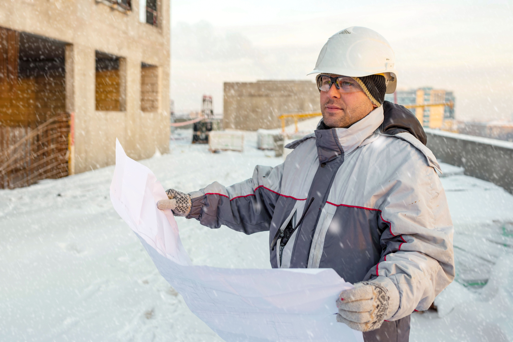 A civil engineer is holding up blueprints while in snowy weather, determining how they'd want to use their concrete admixtures.